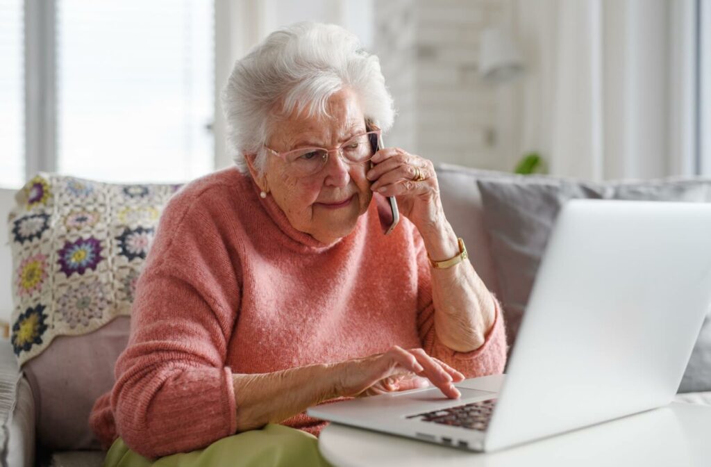 An older adult woman using a laptop and a smartphone while on her couch.