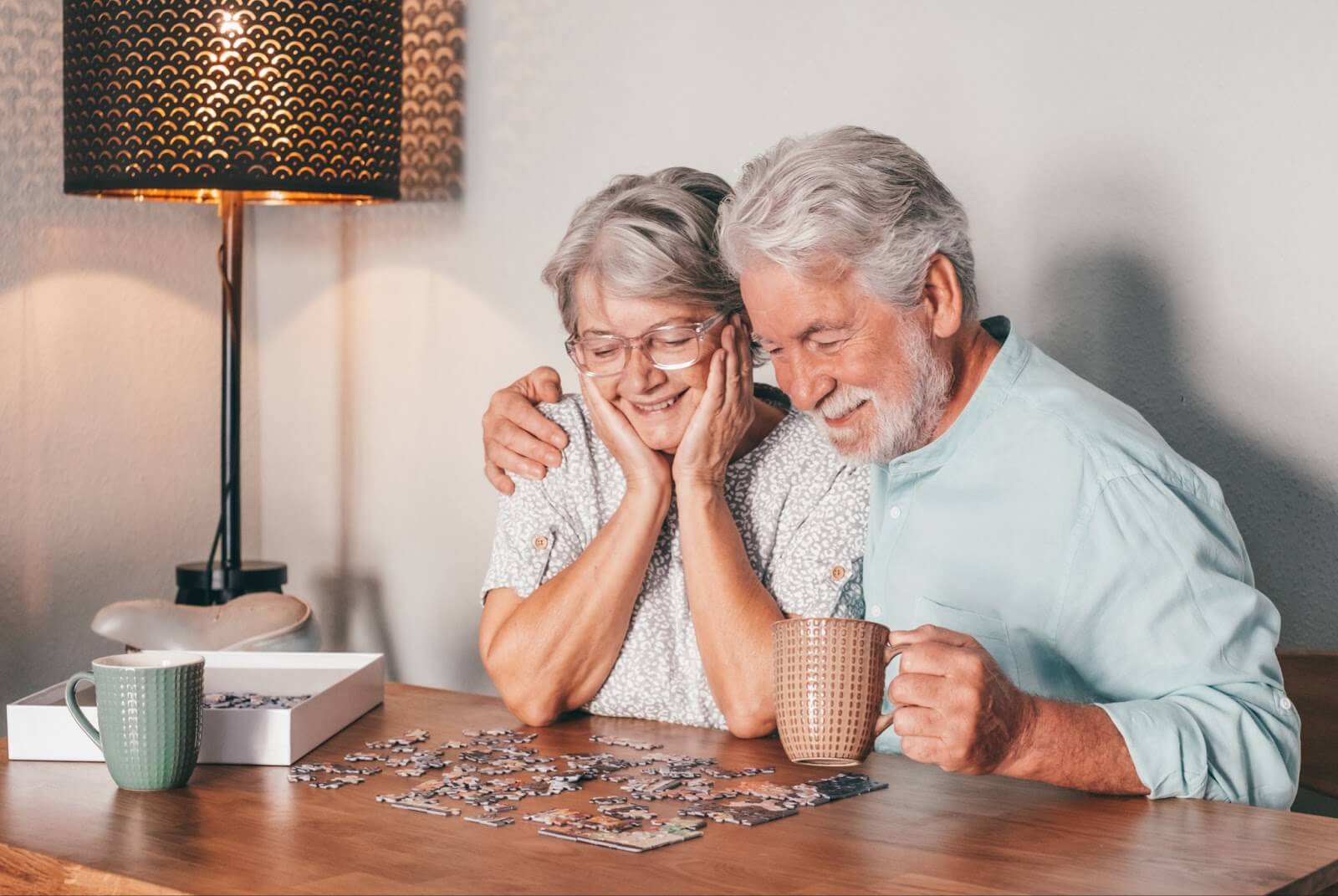 : An older adult couple smiles and enjoys tea while putting together a puzzle.