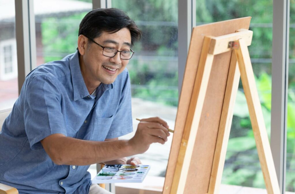 A senior man smiling while he paints, holding a palette with various colors.