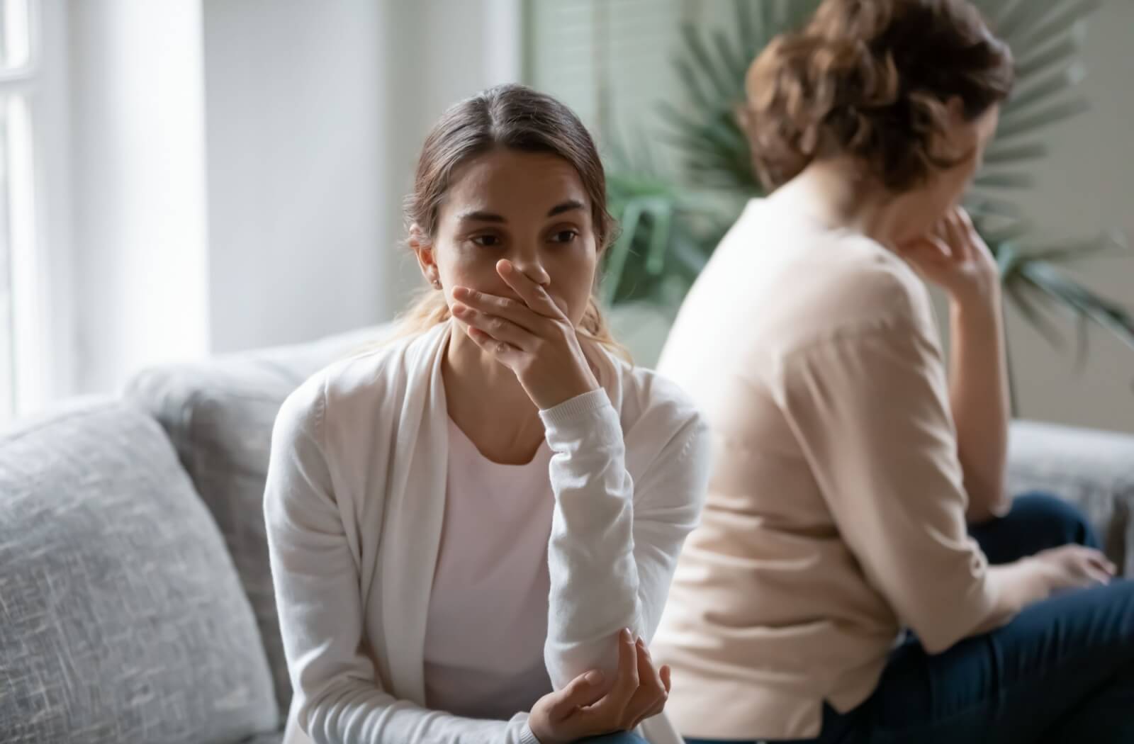 An adult covers their mouth in concern while seated back to back on a couch with their older parent with dementia