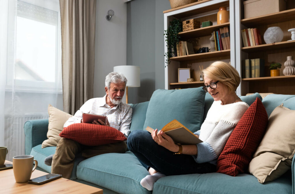 An older couple sitting on their couch in senior living and reading their books together to prepare for their book club.